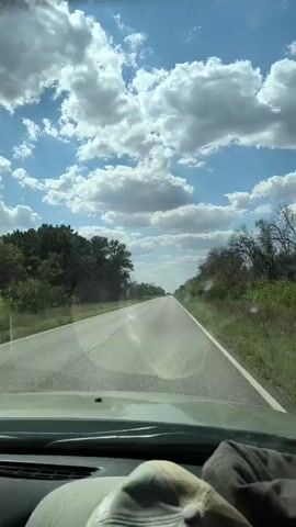 The video shows a view from inside a vehicle, looking out through the windshield at a two-lane road stretching into the distance. The road is lined with green grass and trees on either side. The sky is blue with numerous white, fluffy clouds. A light-colored hat is visible in the foreground on the dashboard. The perspective suggests the vehicle is in motion.