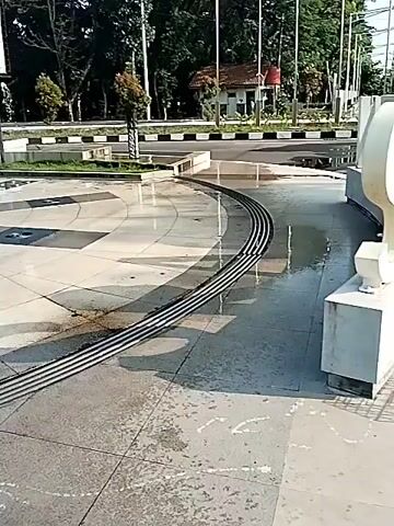 The video shows a public square in what appears to be a city park. At the center is a large statue of a muscular man holding a club. Water flows around the statue and down the plaza. In the background, across a road, there's a monument with curved shapes resembling tusks. There are also small landscaped areas and a small building that might be a guardhouse. The overall impression is of a sunny day at a public space, with the statues as the main feature.