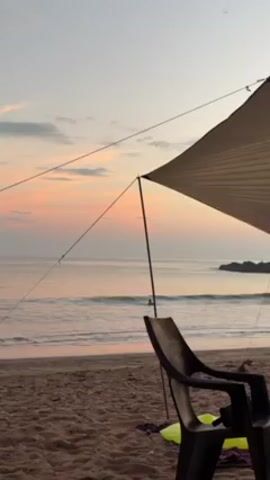The video depicts a beach scene at dusk, with a pastel pink and orange sky visible in the background. In the foreground, a black plastic chair sits on the sand under the shade of a tan-colored canopy supported by thin poles and ropes. The ocean is calm with small waves gently washing ashore. Two individuals are seen entering the water and walking along the shoreline in the distance. The beach appears sandy, and the overall scene suggests a serene and quiet setting.