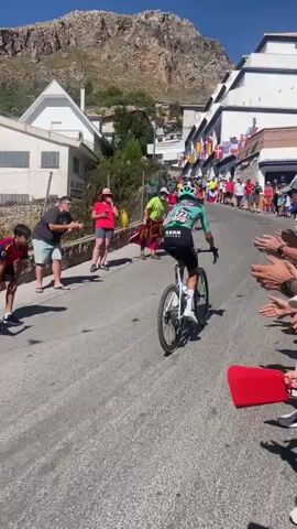 A male cyclist in a teal and black cycling kit with the number 222 on the back is riding a white bicycle up a paved road. He is wearing a green helmet and white shoes. The road is lined with spectators on both sides. On the left side of the road, two young males, one in a red shirt and the other in a grey shirt, are watching the cyclist. Behind them, a man in a grey shirt and shorts is holding his arms up. On the right side of the road, several people are clapping and cheering. In the background, there are white buildings with red-tiled roofs, a steep, rocky mountain, and numerous flags hanging from the buildings. The sky is clear and blue. The overall impression is of a cycling race or event taking place in a town with a mountainous backdrop.
