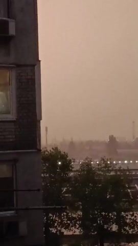 The video captures a hazy, overcast sky, likely during the daytime given the ambient light, though the thick haze obscures the sun. The scene is a view from a building, likely an apartment complex, looking out over a somewhat industrial or urban area. In the foreground, a portion of a building's exterior is visible on the left, with a window reflecting some light and an air conditioning unit mounted on the wall.

Beyond the immediate building, a dense cluster of trees with dark green foliage dominates the mid-ground. The trees are partially obscuring a cityscape in the distance. Visible within the cityscape are several tall, slender structures, possibly communication towers or industrial chimneys, as well as lower-lying buildings. Numerous small, bright lights are scattered across the distance, suggesting streetlights or illuminated windows, indicating the presence of a populated area. The overall atmosphere is muted and somewhat oppressive due to the heavy haze, which gives the sky and the distant landscape a uniform, brownish-pink hue. No people are visible, and the activity appears to be the static view of a hazy urban landscape. The location cues suggest Boyarka, Ukraine. There is no visible text in the media.
