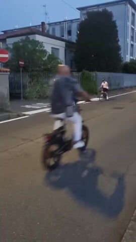 The video opens with a street scene as dusk begins to fall. In the foreground, the asphalt road shows signs of wear with cracks and a white painted line. To the left, a sidewalk and a metal fence are visible, along with some greenery. On the right, another sidewalk borders the road, also with some vegetation. Several people are present. Two young individuals on bicycles appear to be interacting on the road. Further down the street, another person is riding a bicycle, and a fourth person is riding a moped or scooter. In the background, buildings stand adjacent to the road, one tall and modern with multiple floors, and another lower, older structure. The lighting suggests twilight, with streetlights beginning to illuminate the scene, and the headlights of an approaching vehicle are visible in the distance. The overall atmosphere is calm and everyday.