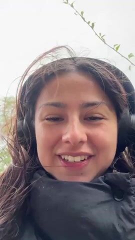 A woman with long brown hair is walking in the rain. She is wearing a black puffer jacket and headphones. She is looking at the camera and talking. The background shows trees and a street with cars. It is a cloudy day.