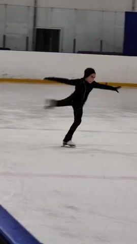 A young figure skater in a black outfit spins on an ice rink. The rink has yellow markings and a white wall in the background with black accents and a door. The skater is in mid-spin, with arms and legs extended. The camera is low, capturing the skater from the waist down.