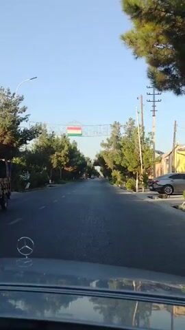 The video shows a view from a car driving down a street lined with trees. An archway is visible spanning the road, decorated with a flag and an ornate design. The sky is clear and blue, suggesting a sunny day. The road surface appears to be asphalt, and there are other cars visible in the distance. The car's hood and a Mercedes-Benz emblem are in the foreground. The overall scene depicts a pleasant, urban or suburban environment.