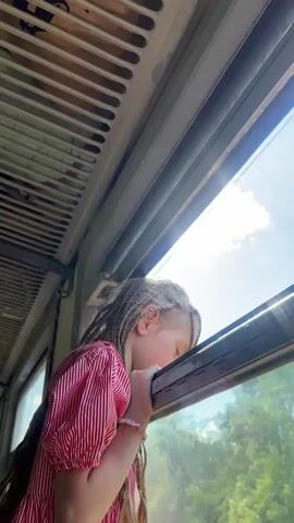 A young girl with braided blonde hair leans her head on a train window, looking out at the scenery. She is wearing a red and white striped dress and a pink bracelet. The sun is shining, casting a bright light through the window. The interior of the train car is visible, with a slatted ceiling and metal framing around the window. The girl's expression appears to be one of quiet contemplation or wonder as she observes the passing landscape. The scene suggests a journey, likely during the daytime, with clear skies. There is no visible text in the media file. The location context provided is Жабінкаўскі сельскі Савет, Belarus.