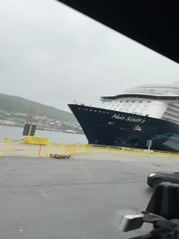 A large cruise ship, named "Mein Schiff 3," is docked at a port in Bergen, Norway, on an overcast day. The ship's dark blue hull and white superstructure dominate the left side of the frame, with its bow pointing towards the viewer. The text "Mein Schiff 3" is prominently displayed on the bow, along with "Genuss" and "Seereise" further down. Yellow safety barriers line the edge of the dock in front of the ship. In the background, green, tree-covered hills or mountains are visible, along with buildings of a town or city nestled along the waterfront. The asphalt ground of the dock appears wet, suggesting recent rain. A crane is also visible in the distance to the left. A black car is partially visible on the right side of the frame, with parts of a car interior (likely the dashboard or headrest) visible in the foreground, suggesting the video was taken from inside a vehicle. The sky is a uniform light gray.