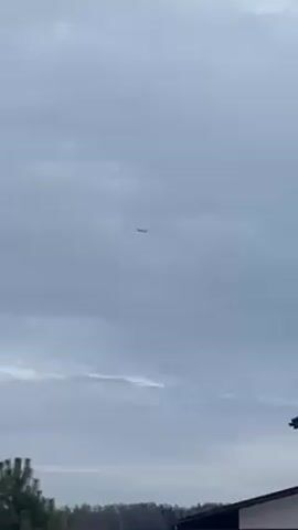The video shows an airplane flying in the cloudy sky above a suburban area. The camera pans up, following the plane, before focusing on the sky and neighborhood. There are houses with dark roofs, trees, and power lines visible. The overall atmosphere is overcast.