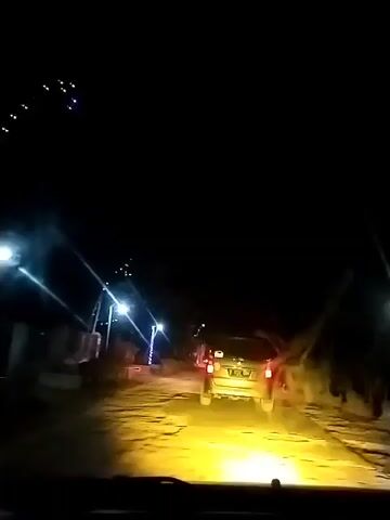 The video begins from inside a vehicle at night, looking out through the windshield. The headlights of the vehicle illuminate a road with a yellow hue, revealing cracks and uneven patches on the surface. To the left and right of the road, there are streetlights that cast beams of light into the darkness. In the distance, a white SUV is visible, its taillights glowing red. As the video progresses, the SUV continues to drive away. The surrounding environment appears to be a rural or semi-rural area, with some vegetation and structures dimly visible in the background. The sky is dark, with a few scattered lights that might be stars or distant buildings. The road appears to be winding slightly. The vehicle the footage is being filmed from is moving, indicated by the slight blur and the changing perspective. The scene depicts a nighttime drive through a sparsely lit road.