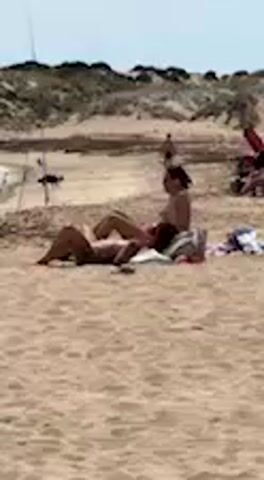 Two people are seen relaxing on a sandy beach during the day. One person lies prone on a striped towel, while another sits upright, looking down at an object in their hands. The seated individual occasionally gestures with their left hand and turns their head slightly. In the background, sand dunes with sparse vegetation are visible, and a small dark animal, possibly a dog, walks along the dune. Other distant figures and beach equipment are faintly discernible on the beach. The sky is bright and clear, indicating sunny weather. The scene depicts a tranquil beach environment, consistent with a location like Barbate, Spain. There is no visible text.