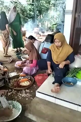 A group of women are gathered outdoors, preparing food. They are sitting on the ground, surrounded by bowls of vegetables and ingredients. Some of the women are chopping carrots, while others appear to be grating something. In the background, a pot is simmering over a fire, indicating that cooking is underway. A motorcycle is parked nearby. The scene suggests a communal food preparation event, possibly for a gathering or celebration.