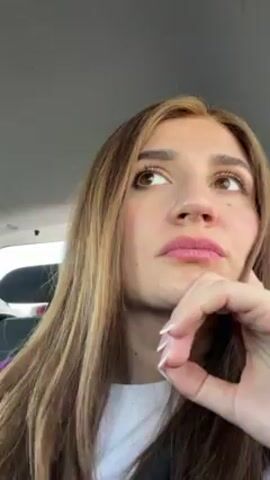 A young woman is in a car, looking pensive. She has long, light brown hair and is wearing a white shirt and black jacket. Her right hand is raised to her chin with a closed fist. She looks up and to the side, then closes her eyes. The car interior is visible, and the ceiling is light gray.