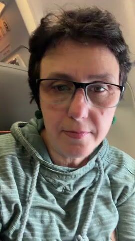 A woman with short, dark hair and glasses is sitting on an airplane. She is wearing a green hooded sweatshirt. She is looking at the camera and speaking. The seat in front of her is visible.