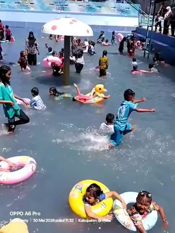 The video shows a shallow, light-blue water pool, crowded with children and a few adults. Most of the kids are either swimming or floating with the help of inflatable rings, toys, or floats. One girl stands in the water, while another lies on a duck-shaped inflatable. A large, mushroom-shaped structure stands in the center of the pool. A line of adults stand along one side of the pool. The video was taken using an OPPO A6 Pro.