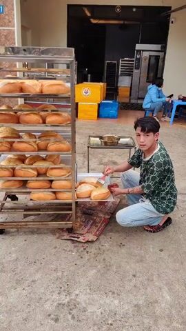 A man kneels on a rug, brushing bread with a pastry brush outside a bakery, where a metal rack is filled with loaves of bread. The man has tattoos on his lower arms and is wearing blue jeans, a green and black printed shirt, and sandals. He dips the brush into a small gold bowl on a metal side table and brushes each loaf. In the background, two people sit on plastic chairs, one using a phone, with yellow boxes stacked behind them. The building number "721" is visible above the door. The ground is concrete. An oven stands in the background.