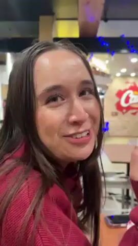 The video shows a young woman singing to the camera. She is wearing a red sweater, and has long brown hair. She smiles a lot and sings, “All I want for Christmas is you.” The camera moves to show a young man next to her, also wearing a red sweater. They are inside what appears to be a restaurant. The restaurant has blue Christmas lights. One restaurant sign says, “WHAT ARE YA…” The man is holding a ring on his ring finger. The man and woman face the camera and interact.