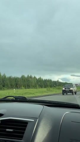 The view from inside a car driving on a highway, in cloudy weather with raindrops on the windshield. There are several vehicles on the road, including a gray pickup truck, a white SUV, a silver sedan, and a yellow sedan. The highway has green fields and trees on either side. The car's dashboard is visible at the bottom of the frame.
