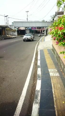 The video shows a view from a sidewalk looking towards a street and an overpass. On the sidewalk, there are tactile paving blocks, a white stripe, and a brick wall with pink flowers. The street is paved and has a white line marking the edge of the lane. A silver car is driving on the street, and a few motorcycles are in the background. An overpass spans the street in the distance. The environment appears to be an urban or suburban area with buildings and utility poles visible. The lighting suggests it is daytime.