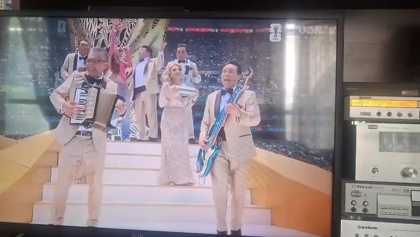 The video shows a live broadcast of a sporting event, likely a soccer match, on a television screen. In the foreground, a band performs on a stage, with members playing an accordion, a bass guitar, and other instruments. They are dressed in matching beige suits. Behind them, a crowd of people in colorful outfits are dancing. The background shows a stadium filled with spectators, a large trophy in the center of the field, and fireworks. The broadcast is labeled with "TUDN en Vivo".