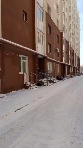 The camera pans across a snow-covered street lined with tall apartment buildings. The buildings are a mix of brown and beige, and red and beige brick. Cars are parked along the street, and a few are driving. There is a decorated Christmas tree on the side of the road, next to a playground. The sky is blue with some clouds.