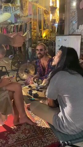 The video shows four people sitting on a rug in a cluttered indoor space. One person is taking a photo of the group with their phone. The rug is ornate and colorful. One person is wearing a grey t-shirt, another is wearing a colorful robe, and another is wearing a light colored dress. One person is partially cropped out of the frame. A fan is visible behind them, as are racks of clothing and various decorative objects. The lighting is dim. The group appears to be engaged in conversation. There are small cups and containers on the rug in front of them.
