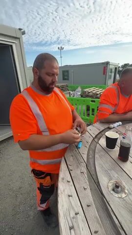 The video shows a group of men taking a break at a construction site. Two men in orange high-visibility vests and pants are sitting at a wooden table. One man is lighting a cigarette. The other man is looking at his phone. Several disposable coffee cups, a can of soda, and a bottle of beverage are on the table, along with a pack of cigarettes and a blue plastic bag. The background shows various industrial containers and fencing, with a cloudy sky above. Another man in a grey t-shirt and a third person, partially visible, are also at the table. The table appears to be made from a large wooden cable spool. The overall atmosphere is casual, suggesting a break from work.
