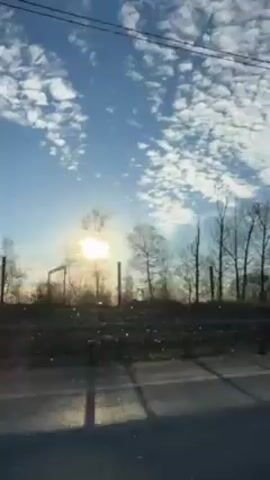 The video shows a view from inside a moving vehicle, likely a car or truck, looking out the passenger-side window. The sun is visible in the distance, low on the horizon and slightly obscured by bare trees. The sky is blue with scattered, puffy white clouds. A grassy or weedy field is visible beyond the road and a fence. Utility lines stretch across the upper part of the frame. The vehicle appears to be traveling on a paved road, indicated by the visible road surface and markings.