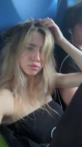 A young woman with long blonde hair is in the foreground of the video, looking at the camera. She is wearing a black tube top and has her hand in her hair. The lighting suggests it is either morning or evening, with sunlight filtering in from the side. In the background, a person with dark hair is visible, also wearing a black shirt. They appear to be in a vehicle, possibly a car or bus, indicated by the blue interior visible in the upper portion of the frame. The overall atmosphere seems casual and intimate, possibly a selfie taken by the woman.