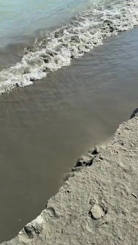 A video shows a close-up of a sandy beach with gentle waves lapping at the shore. The water is a light turquoise color and appears calm. In the background, the landscape is mountainous with green slopes covered in trees. The sky is a clear blue. The video pans out to reveal more of the river and the surrounding scenery. There are no people or objects visible in the video. The overall impression is one of natural beauty and tranquility.