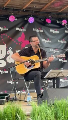 A man is playing an acoustic guitar and singing on a stage. He is wearing a black collared shirt, black pants, and glasses. The stage has a black backdrop with pink butterflies and the words "BÄKGÅRDEN" repeated. There are stage lights above the backdrop. A microphone is positioned in front of the man. A water bottle and a guitar case are visible on the stage. Green grass is in the foreground.