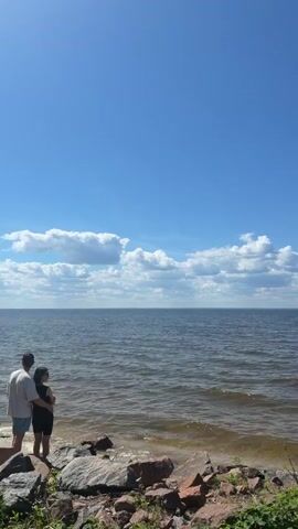 A still, vertical shot shows a body of water under a clear blue sky with scattered clouds. The water is dark and appears to be moving gently, with a brown tint. The foreground is rocky with some green vegetation. There are no people or objects clearly visible in the frame.