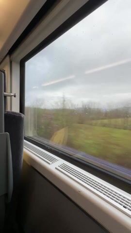 The view outside a train window showcases a countryside scene under a cloudy sky. Raindrops are visible on the glass. Green fields and trees blur by due to the train's motion.