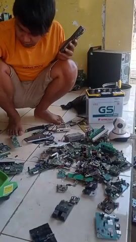 A person wearing an orange shirt and tan shorts kneels on a tiled floor, sorting through a pile of electronic circuit boards. The person holds a small electronic device in their left hand and uses their right hand to sift through the circuit boards. To the right of the circuit boards is a white security camera and a car battery. Behind the person are two computer towers. The environment appears to be outdoors or in an open-air structure, with a yellow wall in the background. Another person's hand is visible on the right side of the frame, reaching towards the circuit boards.