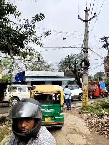 The video shows a street scene with several vehicles and a person. In the foreground, a green and yellow auto-rickshaw is parked on a dirt path. Behind the auto-rickshaw, a white SUV is parked to the left, and a person in a blue shirt is standing near a white car. To the right, a small red truck with a blue tarp is parked, followed by another vehicle. Electrical poles with numerous wires are visible throughout the scene. The environment appears to be a somewhat cluttered urban or semi-urban area with trees, foliage, and buildings in the background. The sky is overcast.