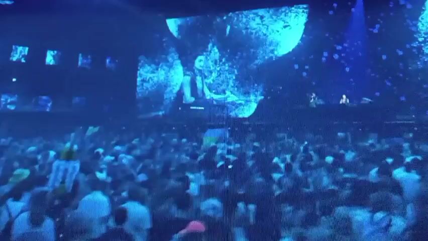 This is a video recording of a live performance. The scene is a concert stage with a large, bright blue LED screen as the backdrop. On the screen, abstract, fragmented images are displayed, mostly in shades of blue and white.

In the foreground, two individuals are visible behind DJ equipment. On the left, a person with short, light-colored hair is operating the equipment. They are wearing dark clothing and appear to be focused on their task. To the right of this person, a woman with long, dark hair and headphones on is standing. She is wearing a shimmering, silver-patterned top and looks engaged with the performance, possibly singing or interacting with the audience. The DJ equipment itself includes a Pioneer brand mixer.

The lighting on stage is predominantly blue, casting a cool and energetic atmosphere. The overall mood seems to be one of excitement and vibrant musical performance. The recording is likely from a night-time event given the stage lighting and the nature of a concert. No specific weather conditions are discernible from the indoor or stage setting. The location context provided indicates the event is in Olișcani, Moldova.