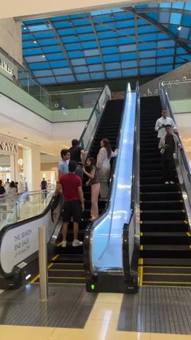 Two escalators are side by side, moving in opposite directions. The left escalator is moving upwards, and the right escalator is moving downwards. Several people are on the escalators. On the left escalator, a group of people, including men and women, are ascending. On the right escalator, two men are descending. Both escalators have black steps and handrails. Above the escalators, a blue ceiling with a grid-like structure is visible. The environment appears to be a shopping mall, with store fronts and other shoppers visible in the background on different levels. There are signs on the sides of the escalators, one of which reads "THE SEASON END SALE."