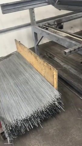 The video shows an industrial setting, likely a factory in Osogóra, Poland. The primary focus is on a wire mesh production machine. The machine is a large, automated apparatus with various metal components, rollers, and a control panel with buttons. It appears to be in operation, as evidenced by the movement of some parts and the presence of cut wires scattered on the floor.

In the foreground, a large bundle of thin metal wires is neatly stacked, ready to be fed into the machine. There are also some wooden planks used as dividers or supports. In the background, the machine is a complex structure of metal frames and moving parts. Some of the machine's components are painted gray, while a portion of the top is green. The walls of the setting are plain, industrial-style, with some visible fixtures. The floor is concrete and appears somewhat worn, with debris and scattered wires indicating ongoing work.

There is no visible text in the video that can be easily read. The lighting suggests an indoor environment, likely during daylight hours, typical for a factory. The overall mood is one of industrial activity and manufacturing. There are no people visible in the footage. The activity captured is the operation of a wire mesh manufacturing machine, processing metal wires into a mesh-like structure.