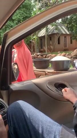 The video shows the interior of a car, looking out the passenger window. The focus is on a woman in a pink hijab standing outside the car, interacting with someone inside. In front of her, on a wooden table, are three large bowls. Two bowls are filled with a white granular substance, likely rice, and one is empty. Beyond the table, a small, rustic building with a tin roof is visible. The surrounding area is lush with greenery, suggesting an outdoor, possibly rural setting. Rocks and debris are scattered around the scene. A hand, presumably belonging to the person filming, is visible on the car door's interior handle, suggesting they are the driver or passenger. The overall impression is that of a transaction or interaction taking place at a roadside stall or a village setting in Nandu, Nigeria. The lighting suggests it is daytime.