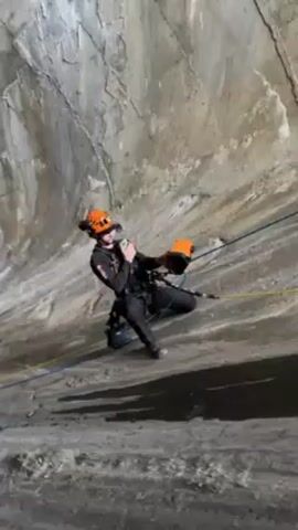 The video shows a man rappelling down the interior of a large, smooth, sloping concrete structure. The man is wearing a safety helmet with lights, a black jumpsuit, and a harness connected to blue and yellow ropes. He is holding what appears to be a measuring device and appears to be communicating with someone using a microphone attached to his helmet. The concrete slope has streaks and stains running downwards, suggesting the presence of moisture or previous water flow. At the top of the slope, there is an opening, likely the exit of the tunnel. The camera follows the man as he descends, maintaining a focus on his movements and the surrounding concrete environment.