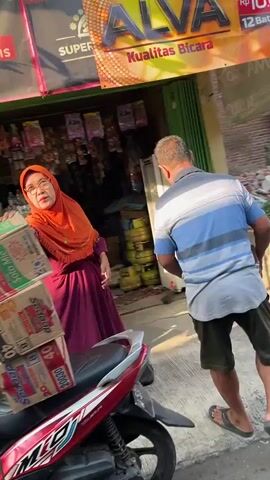 A woman wearing an orange hijab and a purple dress is standing next to a stack of boxes, looking to her left. She is in front of a storefront with a sign that reads "ALVA KUALITAS BICARA." A man wearing a blue and white striped shirt and dark shorts is walking away from the woman, towards the right side of the frame. A red and white motorcycle is parked in the foreground. The scene appears to be an outdoor market or shop.