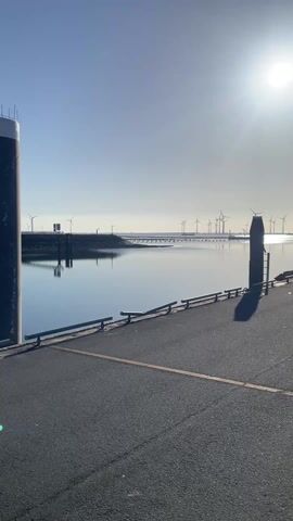 The camera pans across a paved pier on a sunny day. The pier has wooden railings on the left side and is lined with a yellow stripe. In the distance, over a calm expanse of water, are several wind turbines. The sun is bright and high in the sky. As the camera pans right, a few cars and low buildings come into view. The sky is a clear blue.
