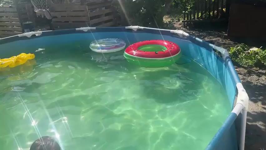 This is a video clip of a young boy splashing in a backyard swimming pool. The boy is the central subject, his face visible as he creates a splash with his hands. He is shirtless, suggesting warm weather. Several inflatable pool toys are visible in the water: a yellow alligator float, a blue inflatable arm float, a clear inflatable ring with colored accents, and a red and green inflatable ring, possibly designed to resemble a watermelon. The pool appears to be an above-ground model with a blue liner.

The setting is outdoors, with lush green foliage and trees surrounding the pool. In the background, there are wooden structures, possibly a fence or a shed made from pallets, indicating a residential backyard. Sunlight is prominent, casting bright reflections on the water and creating a summery atmosphere. The weather appears to be sunny and pleasant.

The activity is a boy enjoying himself, playing and splashing in the pool. His facial expression suggests happiness and exuberance. The time of day is likely daytime, given the strong sunlight. There is no visible text in the clip. The overall impression is one of a carefree summer day spent having fun in the water.