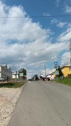 A video shows a rural road with buildings on either side. There are two pedestrian crossing signs visible, one on each side of the road. A tractor with a trailer is stopped in the middle of the road. A white car is parked on the left side of the road. The sky is blue with white clouds. The weather appears to be sunny. The video is shaky, suggesting it was filmed by someone walking or riding a bicycle.