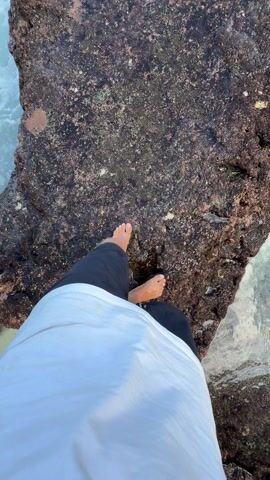 A person stands barefoot on a large, dark, textured rock, wearing black shorts and a white shirt. The camera angle is from the person's perspective, looking down at their feet and then tilting up to reveal the ocean. The ocean water is turquoise and white, with waves crashing against the rocks. In the distance, a green, rocky headland is visible under a clear blue sky. The camera remains stationary throughout the shot.