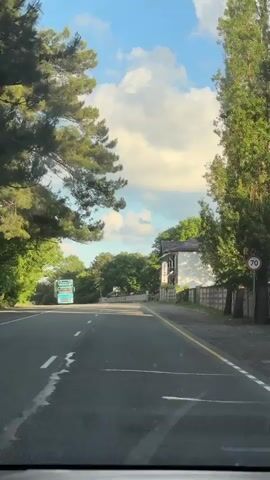 A dashcam view from a vehicle moving on a road on a bright sunny day. The sky is blue with scattered white clouds. On the left side of the road, there are trees and a large blue truck. On the right side of the road, there is a building with a sign that reads "HOTEL ASTORIA". The road is paved and has white lane markings. The vehicle continues to drive forward, passing the building and trees.