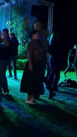 A group of people are dancing outside at night. The scene is illuminated by colored stage lights, casting green and blue hues on the grass. In the foreground, a bald man in a jacket and jeans is dancing with a woman wearing a brown top and a long dark skirt. They are both smiling and appear to be enjoying themselves. Other people are visible in the background, also dancing. A stage structure with lights and speakers is visible in the upper left corner of the frame.