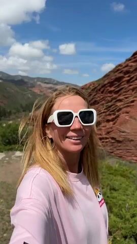 A woman with long, light brown hair and white sunglasses is featured in a close-up selfie video against a stunning natural backdrop. She wears a light pink long-sleeve shirt with a cartoon character, possibly a panda-like figure with a helmet, on the left chest. Throughout the video, she smiles broadly, appearing happy and engaged with her surroundings.

The setting is an arid, mountainous landscape, characterized by dramatic, layered red rock formations that rise steeply in the background. Green shrubs and sparse vegetation dot the foreground and lower slopes of the mountains. The sky is bright blue with scattered white clouds, indicating a clear, sunny day. The woman first appears in profile, looking towards the scenery, then turns to face the camera, capturing herself and the expansive red cliffs of Jeti-Ögüz, Kyrgyzstan.