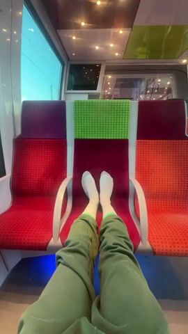 A first-person perspective shot from inside a train or tram shows a person's legs and feet resting on the seat opposite them. The person is wearing green sweatpants and white socks with green accents. The train seats are upholstered in a patterned fabric, primarily red, with accents of maroon, purple, and bright green on the seatbacks. The interior of the train is brightly lit, with ceiling lights visible, and daylight can be seen through the windows, suggesting it is daytime. The scene conveys a sense of relaxation or comfort during a journey, likely in Creil, France, given the provided context.