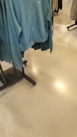 The video shows a retail store, likely a clothing and shoe store. The floor appears to be concrete. The first shot shows racks of clothing, primarily light-colored sweaters or hoodies. A mirror is visible on the left. The second shot pans across more clothing racks, featuring light pink and beige items, as well as black bags. The third shot shows shelves displaying shoes and numerous orange boxes, presumably shoe boxes, stacked high. A person in a blue shirt and shorts is visible in the distance, walking down an aisle. The overall impression is that of a well-stocked retail environment.