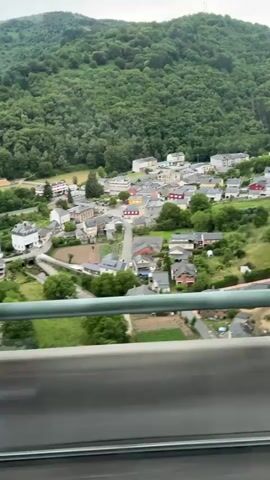The video shows a view of a small village nestled in a valley, surrounded by lush green forested hills. The village consists of numerous houses with pitched roofs, some of which are painted in various colors like yellow and red, interspersed with white and gray buildings. A road winds through the village, with cars visible. In the background, a river or body of water can be seen, with some sandy banks. The foreground is dominated by a railing and some foliage, suggesting the video is filmed from an elevated viewpoint, possibly a train or a vehicle on a bridge. The sky is overcast, indicating a cloudy day. The overall impression is of a serene rural landscape.